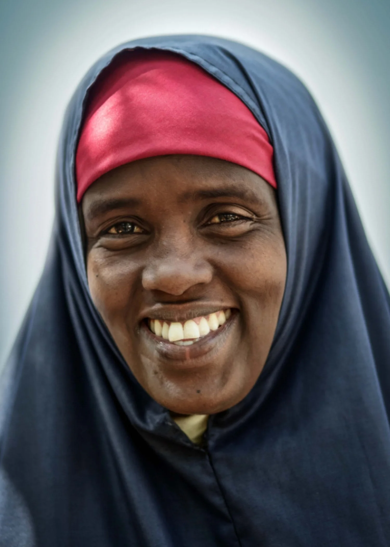 Smiling woman in a red headscarf and grey shawl photographed against a dark backdrop